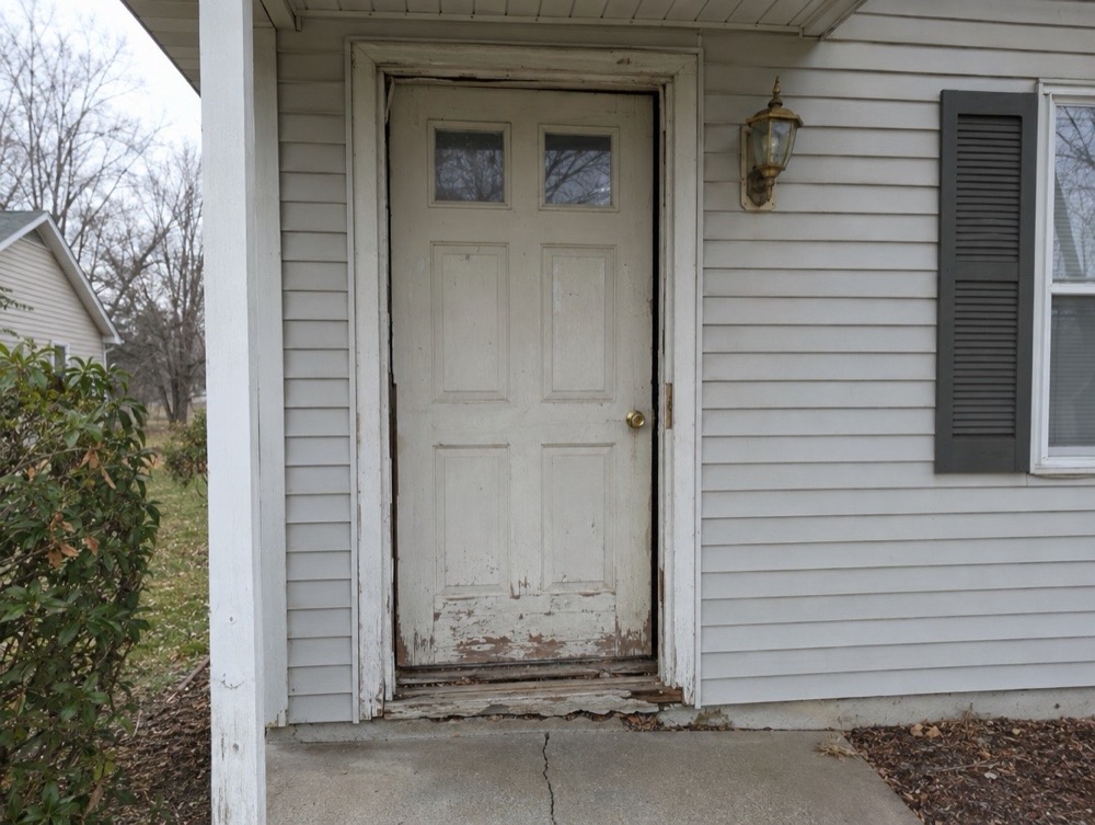 Worn, weathered white front door with a damaged frame and a gap at the threshold