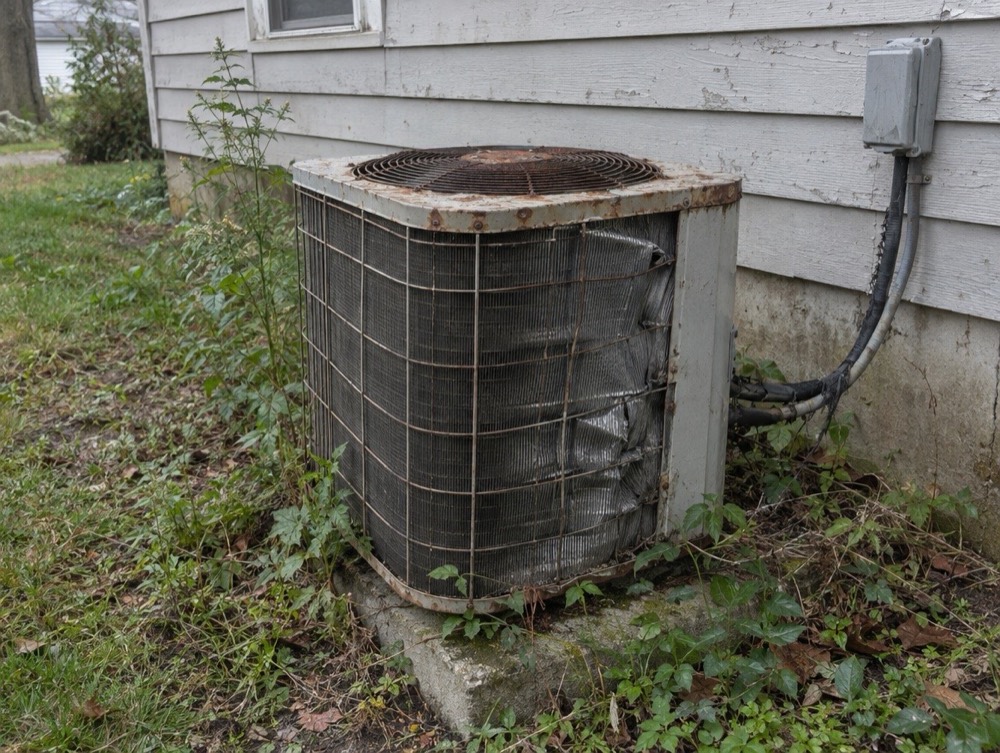 Rusty, weathered air conditioner condenser surrounded by weeds beside a house